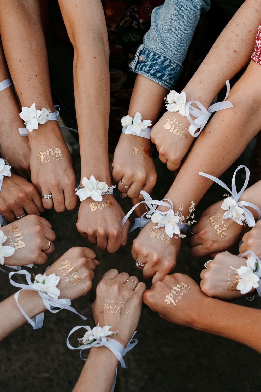 Junggesellenabschied Geschenk: Hände im Kreis mit Blumenarmbändern und goldenen “Team Bride”-Tattoos