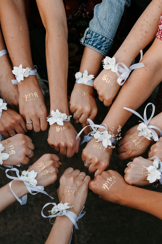 Junggesellenabschied Geschenk: Hände im Kreis mit Blumenarmbändern und goldenen “Team Bride”-Tattoos
